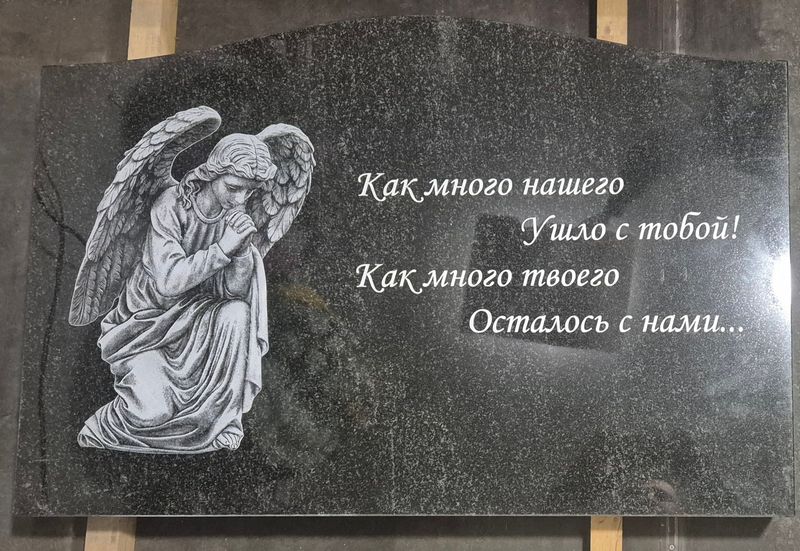 горизонтальная плита из тёмного гранита с гравировкой коленопреклонённого ангела и эпитафией курсивом, стоит на подставке в помещении