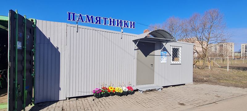 Павильон мастерской крупным планом: объёмная вывеска «ПАМЯТНИКИ», вход с козырьком, ящики с цветами вдоль стены
