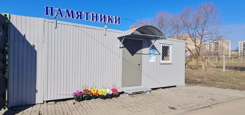 Фасад павильона весной: серые стены, синяя вывеска «ПАМЯТНИКИ», клумбы у двери, брусчатка
